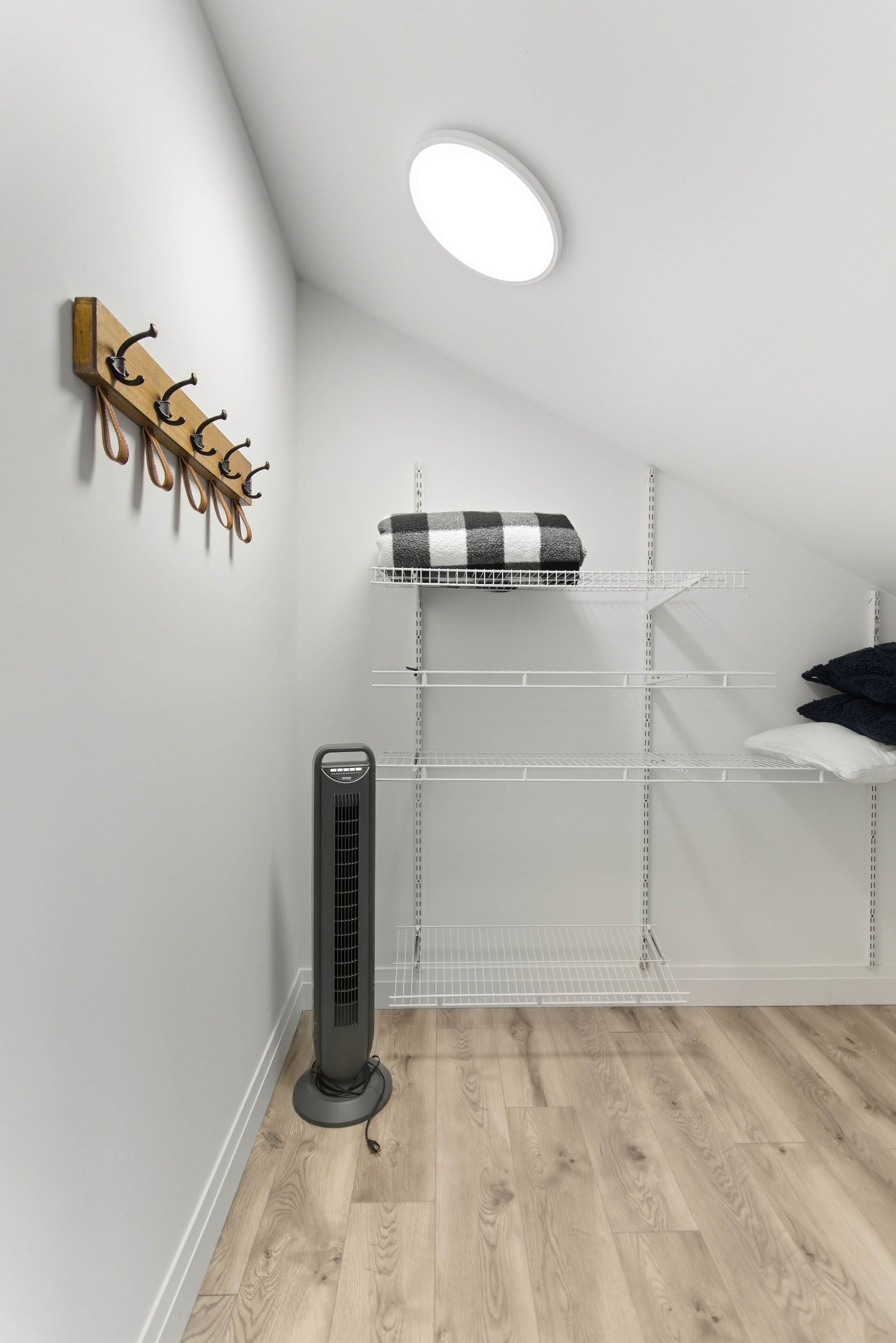 A room with a sloped ceiling, a standing heater, a white shelf unit with a checkered blanket, and a wooden coat rack.