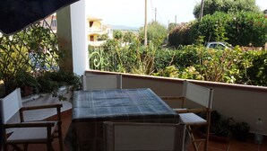 A balcony with a table and chairs, surrounded by greenery and a view of buildings in the distance.