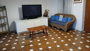 A living room with a television on a white cabinet, a wicker sofa with blue cushions, a wooden bench, and a patterned floor. - Sardinia! Budoni 1 km from the wonderful beach of Sant'Anna: 4 beds (Tanaunella)
