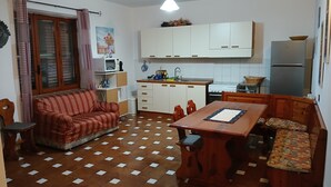 A compact living space with a sofa, a dining table with benches, a kitchen area with cabinets and appliances, and a patterned floor. - Sardinia! Budoni 1 km from the wonderful beach of Sant'Anna: 4 beds (Tanaunella)