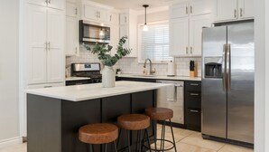 A modern kitchen with white cabinets, a stainless steel refrigerator, a central island with stools, and a hanging light fixture.