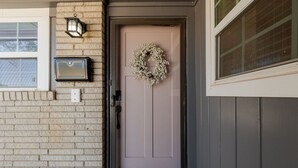 A house entrance with a light purple door, a wreath, a mailbox, and a window with a view of a roof.