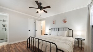 A bedroom with a bed, a ceiling fan, a bedside table with a lamp, and a door leading to another room.