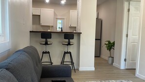 A modern living room with a grey sofa, a white fluffy rug, and a remote control on the sofa. There are two bar stools and a kitchen area in the background. - Airport bungalow  (Coraopolis)