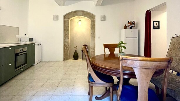 Un comedor con una mesa redonda de madera y sillas, una cocina con armarios verdes, y un arco de piedra que conduce a otra habitación.