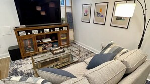 A living room with a sectional sofa, a glass coffee table, a flat-screen TV on a wooden stand, and framed artwork on the wall.