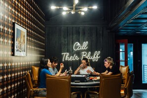 Um grupo de pessoas sentadas em volta de uma mesa em um quarto com parede de madeira escura e um letreiro de neon que diz 'Call Their Bluff'.