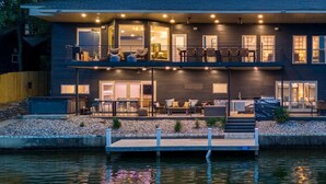 Ein modernes, zweistöckiges Haus mit einer Anlegestelle, die in einen See hineinreicht, Sitzgelegenheiten im Freien und einer gut beleuchteten Terrasse.