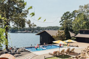 A swimming pool with lounge chairs and umbrellas, a dock, and a view of a lake with houses in the background. - LakeFront with Pool! HotTub! Game Room! Sleeps 20+

 (Hot Springs)