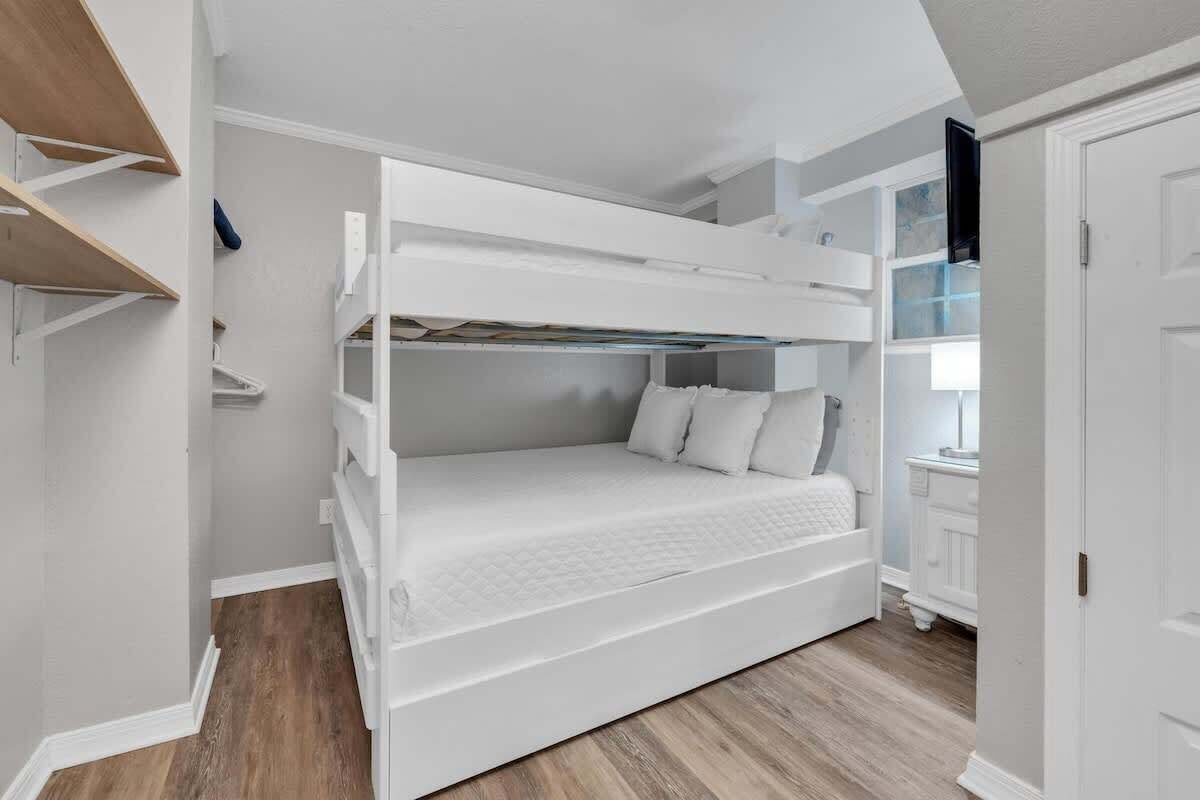 Una cama litera con colchón y almohadas blancas, suelo de madera y una puerta blanca con un televisor encima.