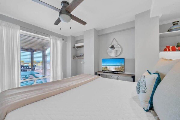 A bedroom with a bed, a ceiling fan, a TV, and a view of the beach through the window.