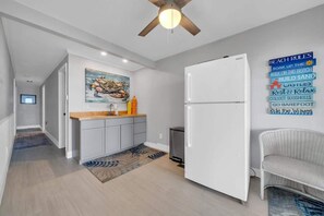 A modern kitchen with a white refrigerator, grey cabinets, and a wooden countertop. There is a ceiling fan, a painting of a boat on the wall, and a rug on the floor.