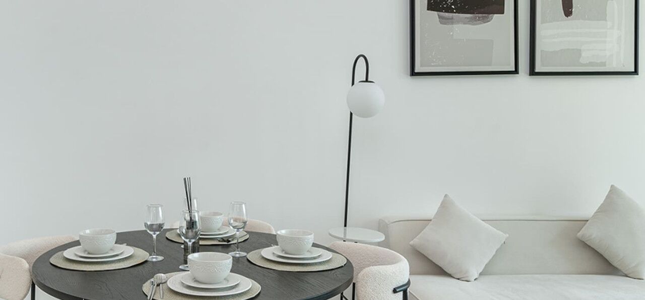 A modern dining area with a round table set for four, white plates, bowls, and glasses, a floor lamp, and a white sofa with cushions.