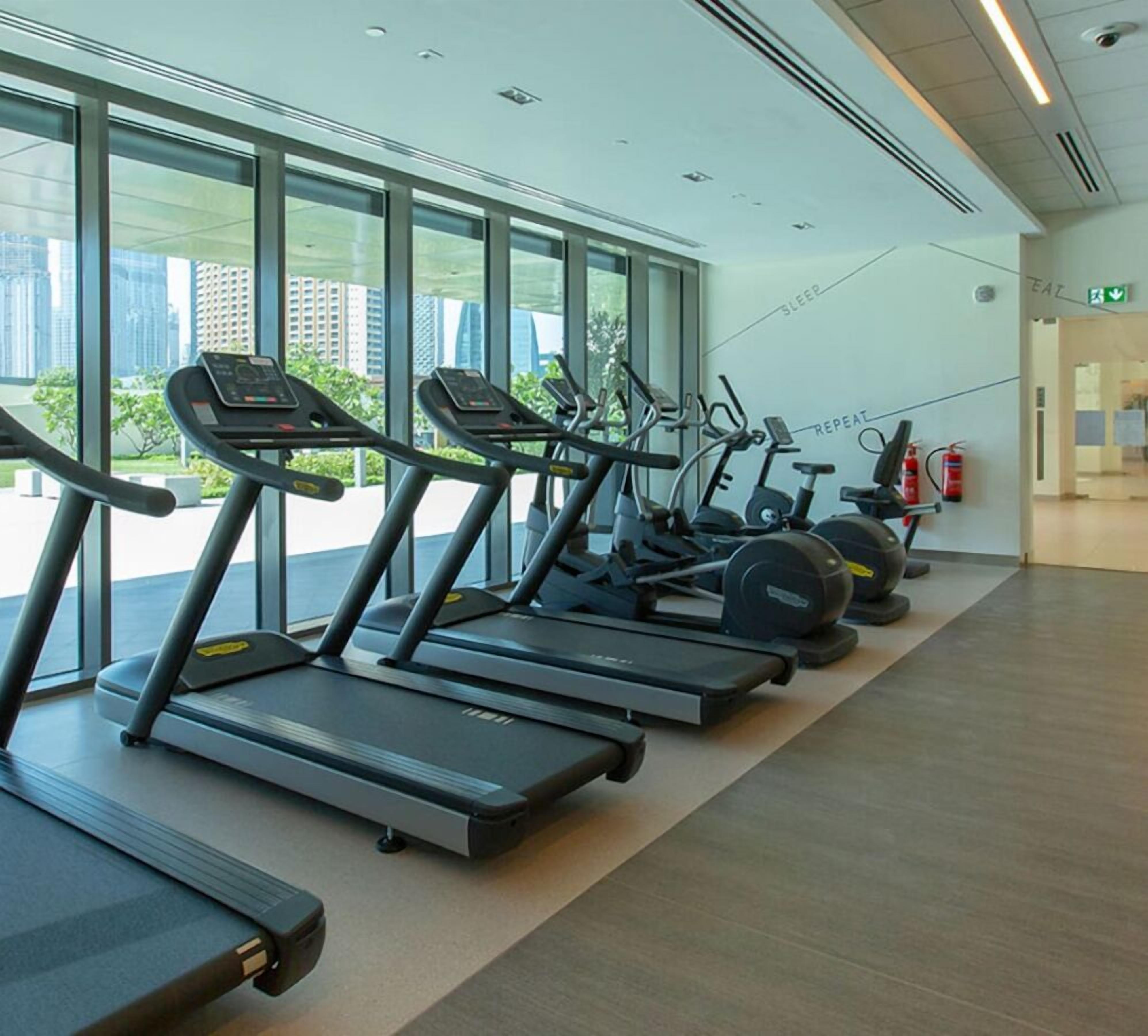 A row of treadmills in a gym with large windows and a fire extinguisher on the wall.