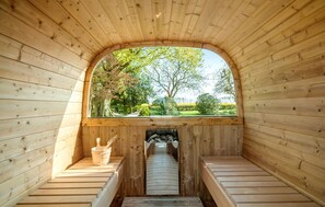 Una sauna con paredes y bancos de madera, una ventana que ofrece vistas a árboles y un jardín, y una chimenea con leña.