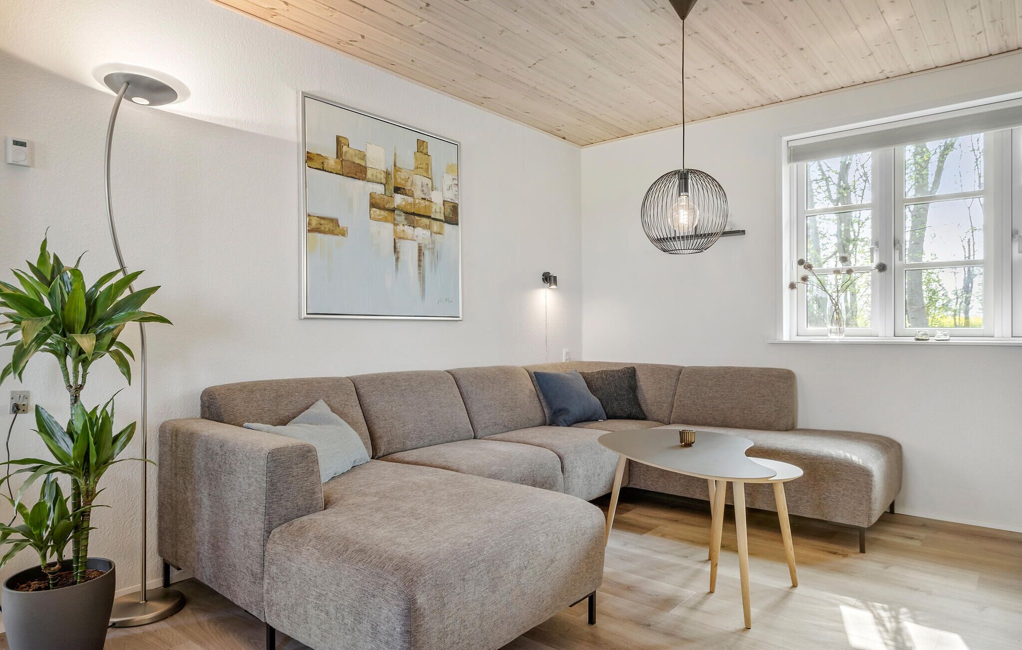 Ein modernes Wohnzimmer mit einem Ecksofa, einem kleinen runden Tisch, einer Topfpflanze, einer Wandleuchte und einem Fenster mit Blick auf Bäume.