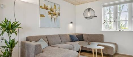 Ein modernes Wohnzimmer mit einem Ecksofa, einem kleinen runden Tisch, einer Topfpflanze, einer Wandleuchte und einem Fenster mit Blick auf Bäume.