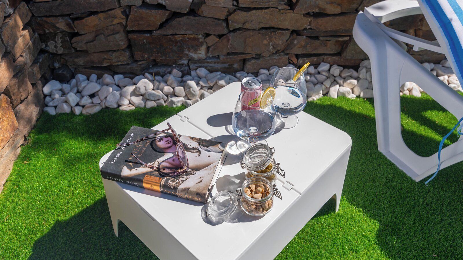 Una mesa blanca con una revista, dos vasos con rodajas de limón, una lata de refresco y frascos de nueces sobre un césped con un muro de piedra al fondo.