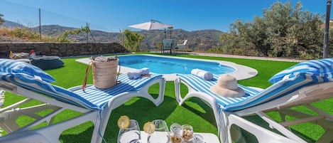 A poolside area with two lounge chairs, a table with drinks and snacks, and a view of a mountain range.