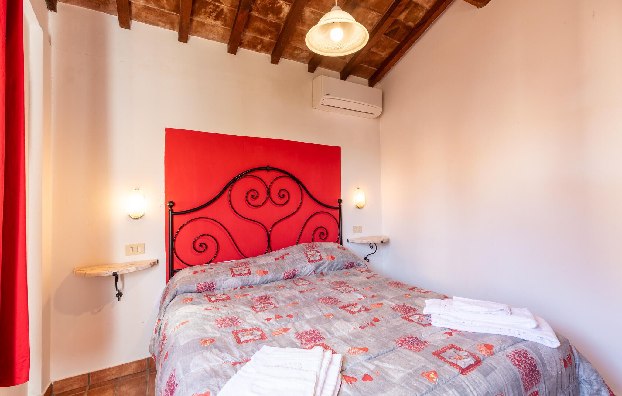 A bedroom with a red headboard, a bed with patterned bedding, two bedside tables with lamps, and an air conditioning unit on the wall.