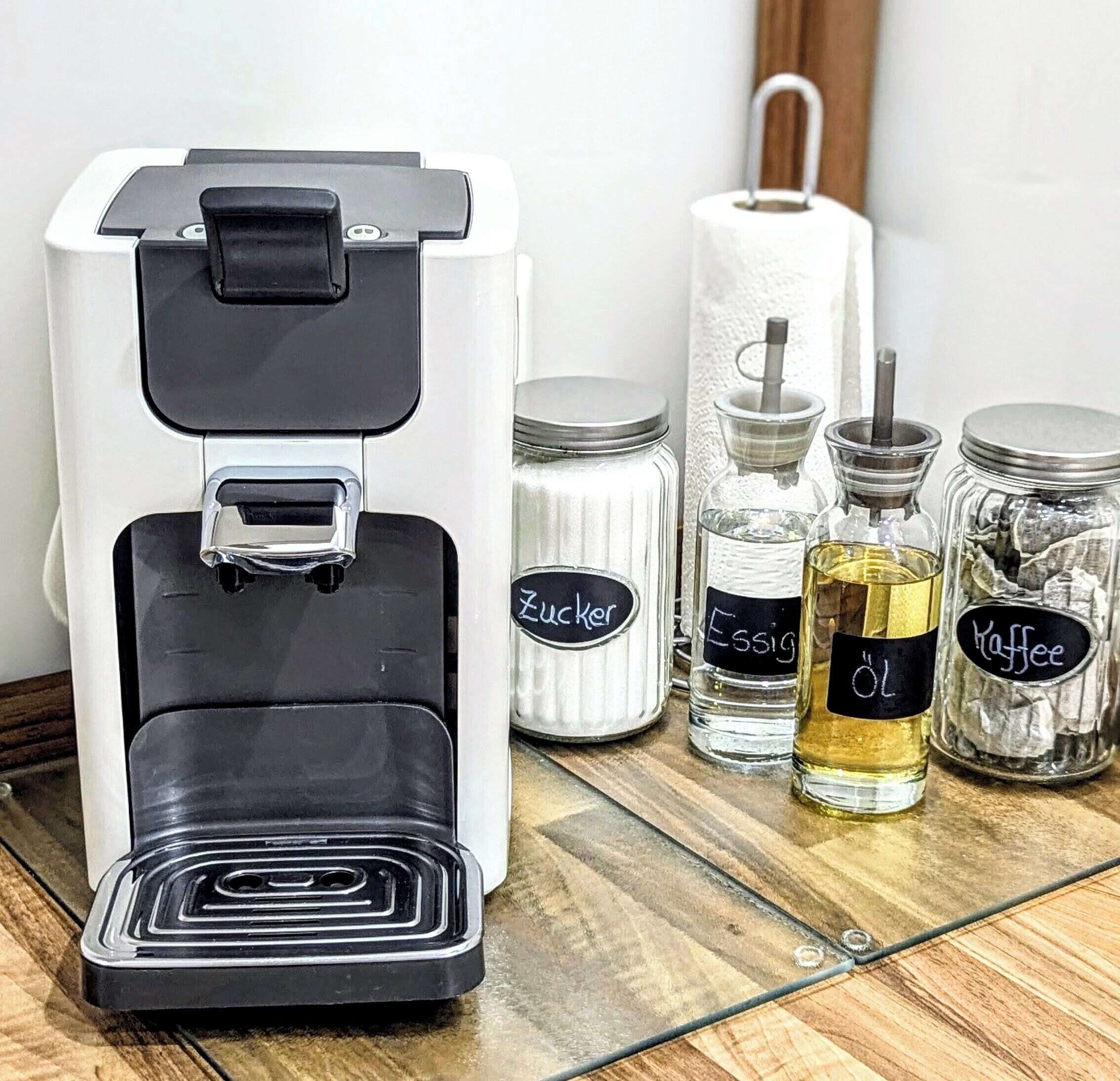 A coffee machine with a black and white design, a roll of paper towels, and three labeled glass jars containing coffee-related items.