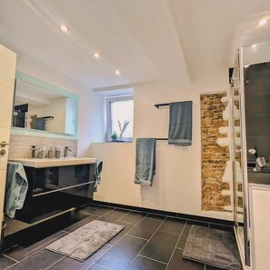 A modern bathroom with a large mirror, dark tiles, a stone accent wall, and a glass shower enclosure.