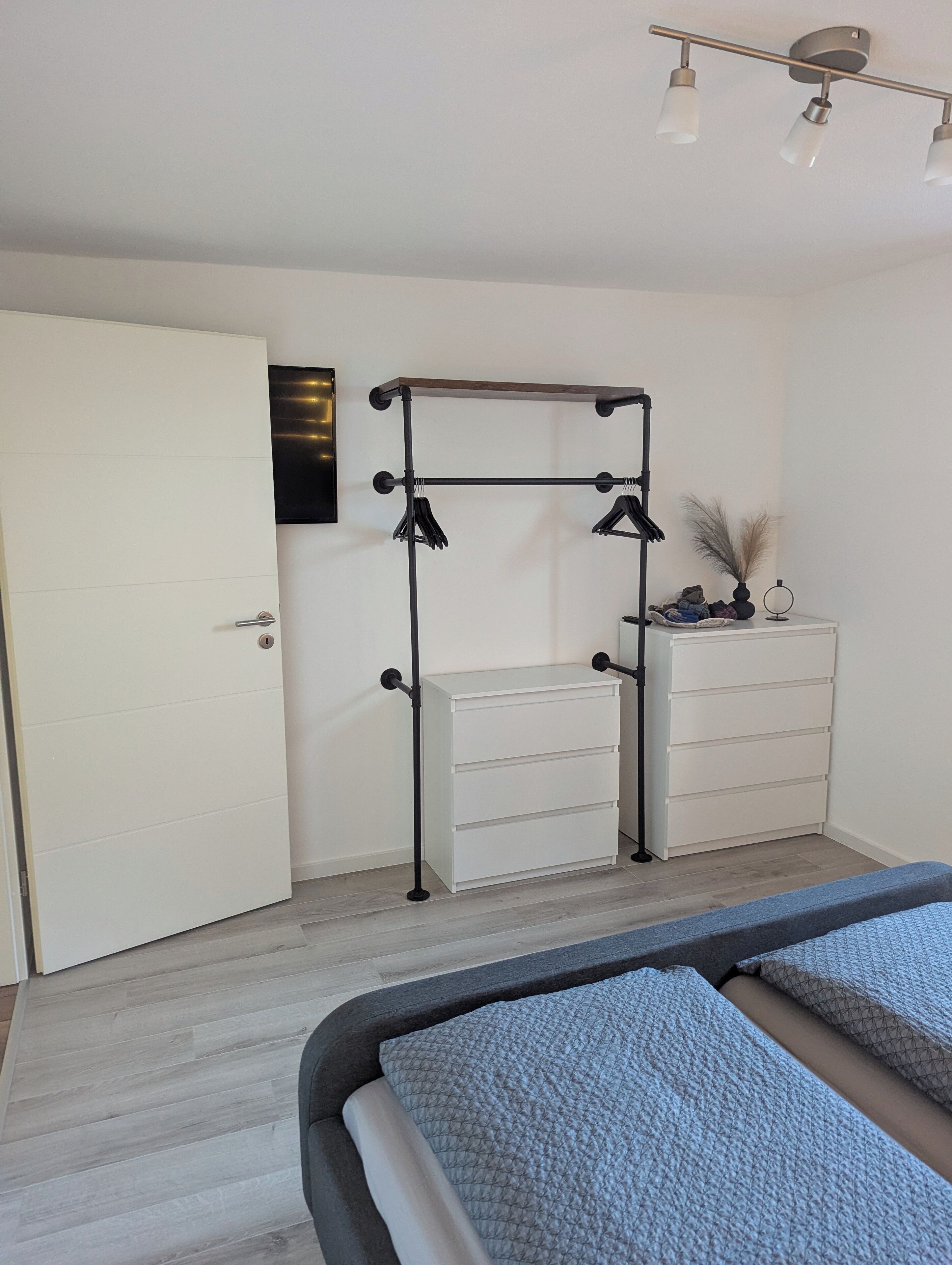 A bedroom with a bed, a white dresser, a black metal shelf unit, and a ceiling light fixture.