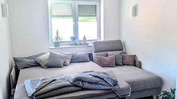 A room with a bed, a sofa, a window with blinds, a potted plant, and a wooden floor.
