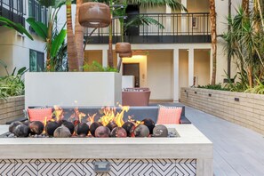 Ein Feuerkorb mit Steinen und eine Sitzecke, umgeben von Pflanzen, mit einem Balkon im Hintergrund.