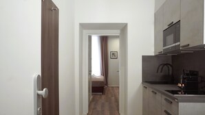 A narrow hallway leading to a bedroom with a bed and a window with curtains. A modern kitchen with a microwave and a sink is visible on the right.