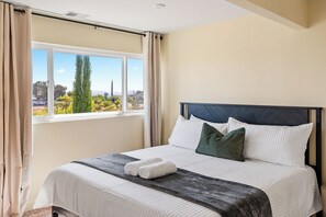 Een net opgemaakt bed met witte lakens en een donkere plaid, een raam met gordijnen, en uitzicht op groen buiten.