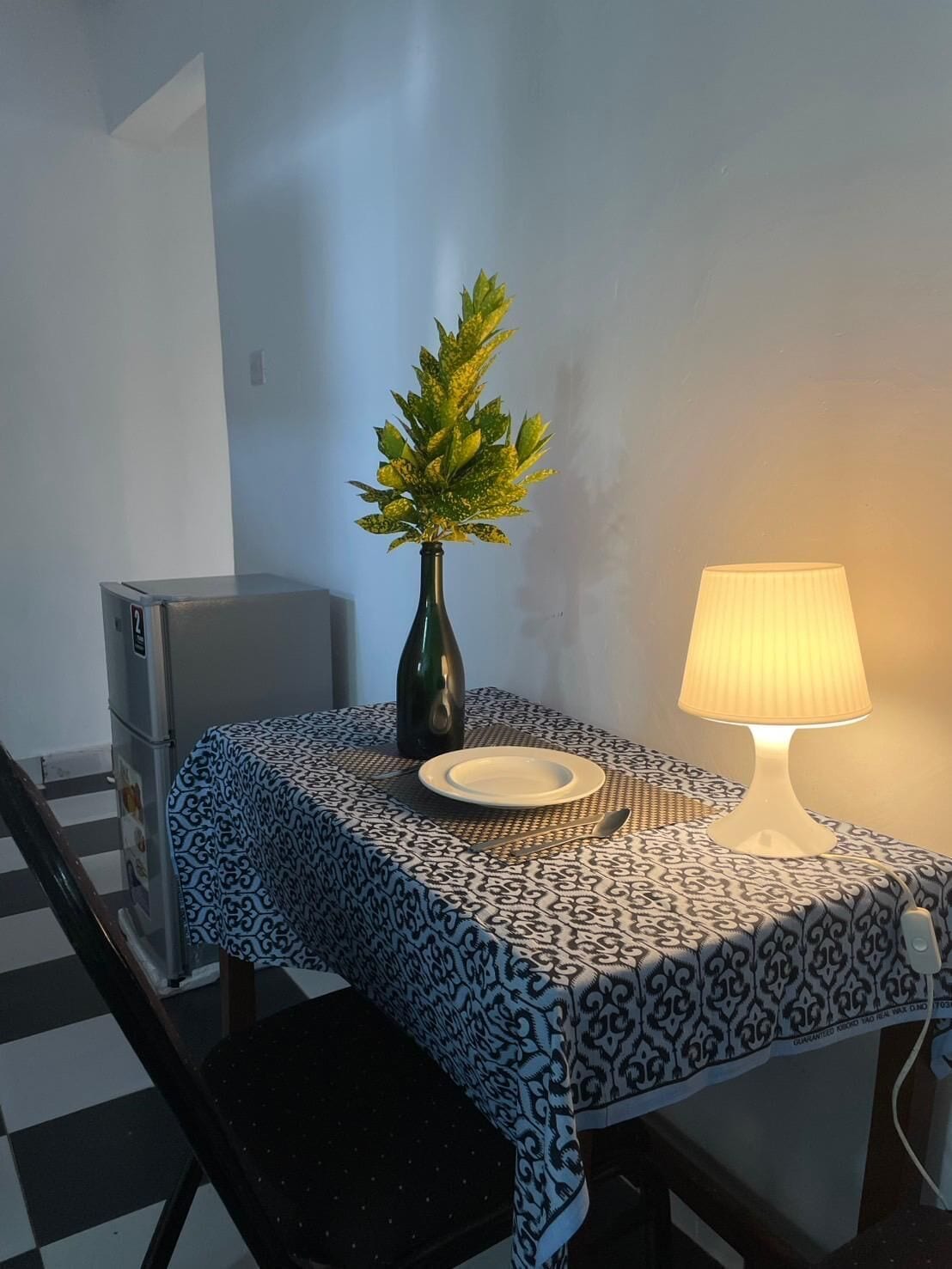 A small dining area with a table covered by a patterned tablecloth, a vase with a plant, a lamp, and a refrigerator.