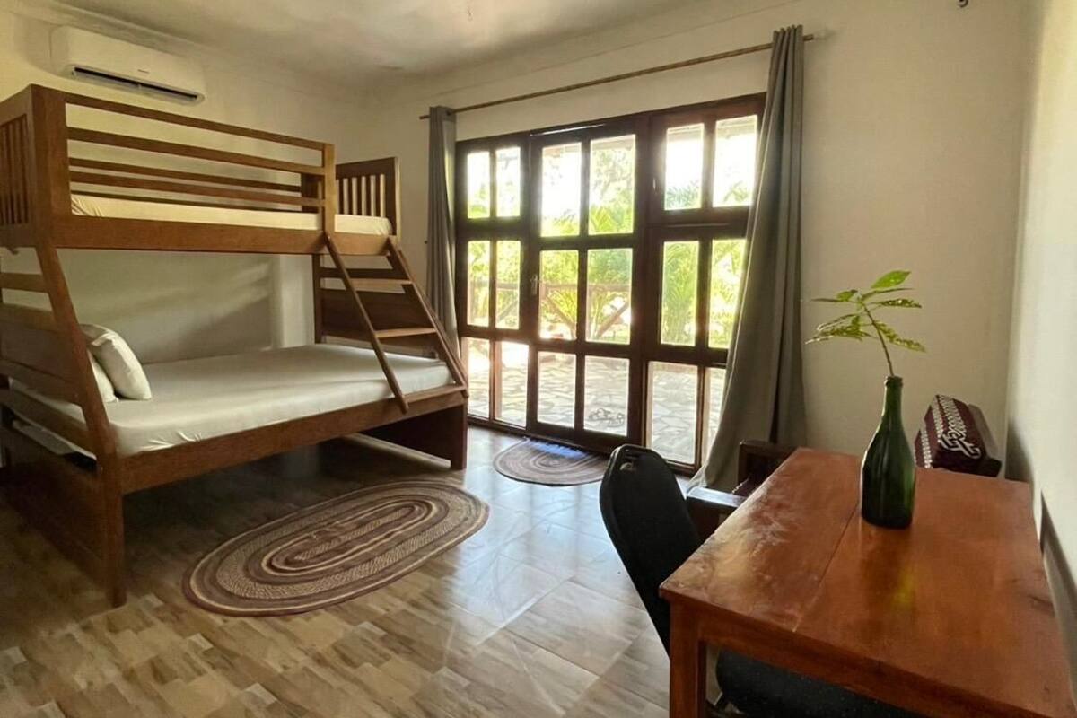 Une pièce avec un lit superposé, une table à manger avec des chaises, un vase avec une plante et de grandes fenêtres avec des rideaux.
