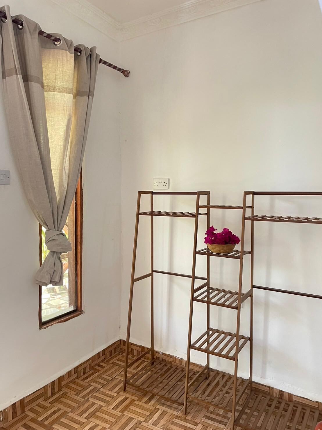 A room with a wooden floor, a window with a curtain, and a metal shelf unit with a basket of flowers.
