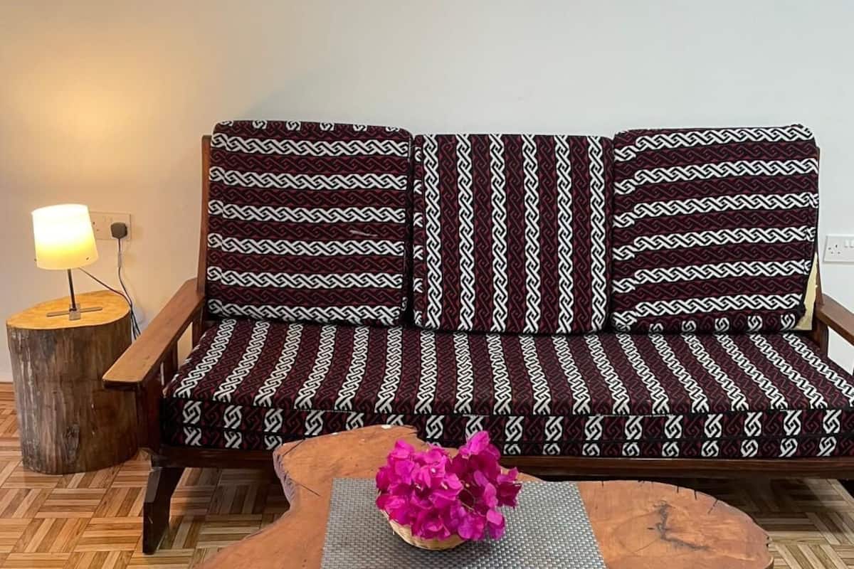 Un canapé en bois avec des coussins à motifs, une table ronde en bois surmontée d’un set de table en fibres tressées, et une lampe posée sur un tabouret en bois.