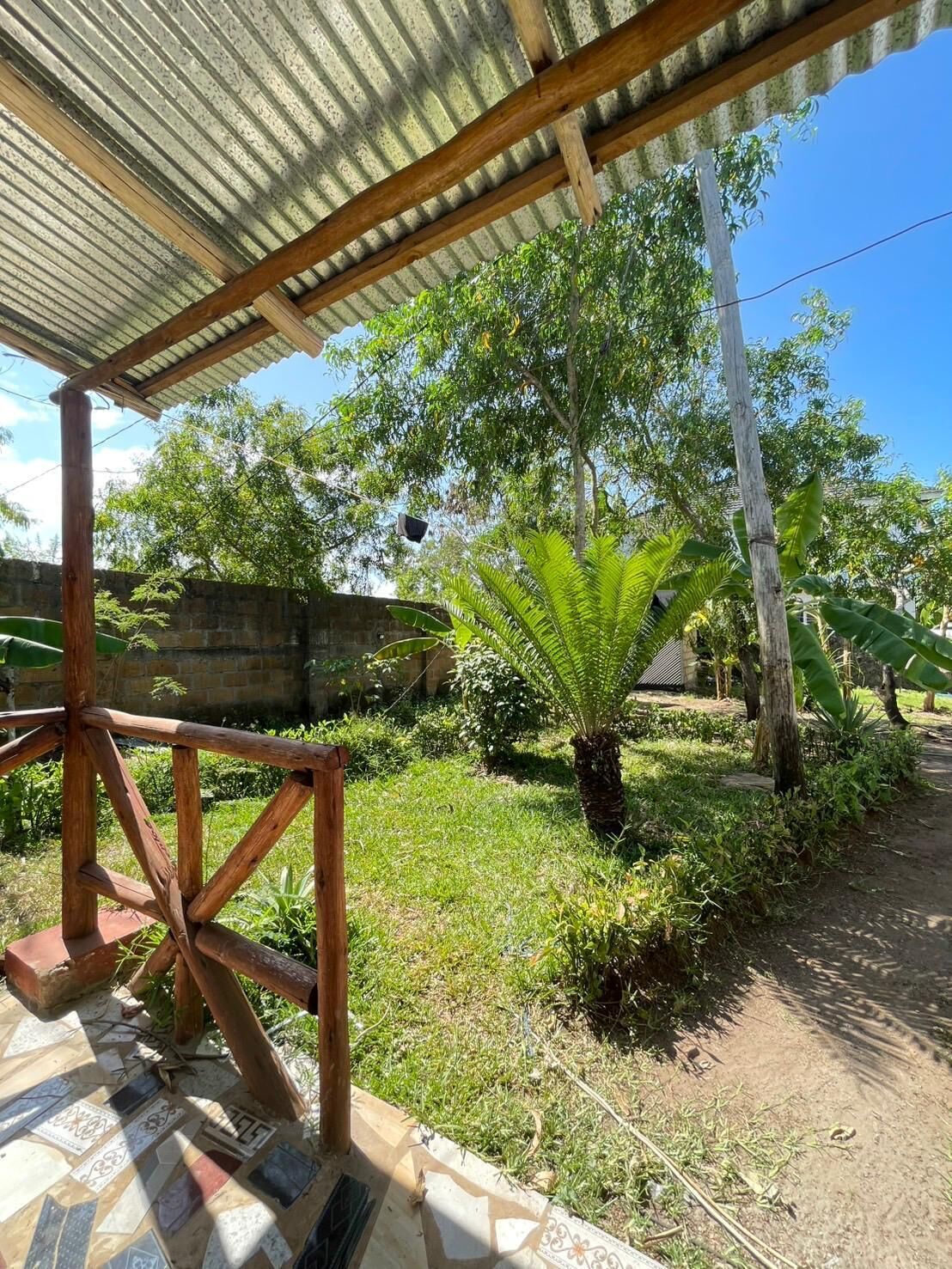 A covered outdoor area with a wooden structure, a stone wall, and a variety of greenery including a fern.