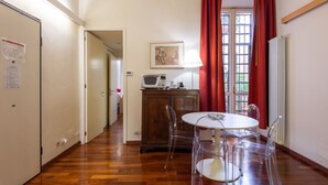 A hotel room with a wooden floor, a round dining table with clear chairs, a microwave, a cabinet, and a window with red curtains.