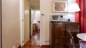 A hotel room with a wooden cabinet, a white table, a clear chair, a framed artwork on the wall, and a hallway leading to another room.