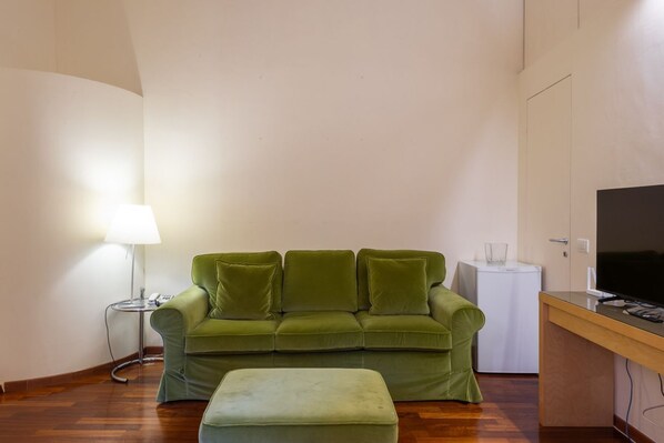 Un canapé vert, une petite table avec une lampe, un téléviseur et un lave-linge dans une pièce dont le sol est recouvert de parquet.