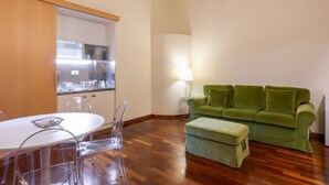A modern living room with a green sofa, a small table with chairs, a lamp, and a kitchen area with cabinets and appliances.
