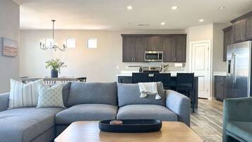 A modern living room with a grey sectional sofa, a wooden coffee table, and a kitchen area with dark cabinets and stainless steel appliances.