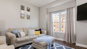 A living room with a beige sofa, a matching ottoman, a blue and white patterned rug, and a large window with curtains.