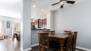 A dining area with a wooden table and chairs, a ceiling fan, and a kitchen in the background.