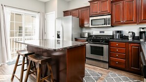 A modern kitchen with a large island, stainless steel appliances, wooden cabinets, and a view to the outside.