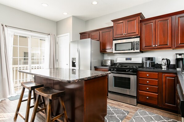 A modern kitchen with a large island, stainless steel appliances, wooden cabinets, and a view to the outside.