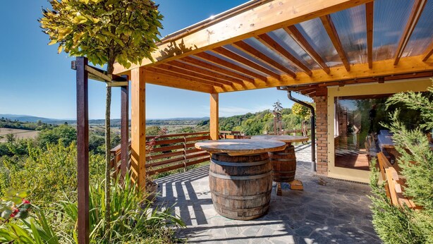 Une pergola en bois installée à l’extérieur, avec un sol en pierre, une table en bois et un tonneau.
