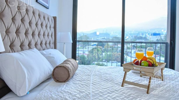 A bedroom with a large bed, a view of a cityscape, and a tray of fruit and juice on the bed.