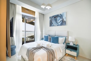 A modern hotel room with a large bed, a view of the city through floor-to-ceiling windows, a painting on the wall, and a nightstand with a lamp. - Exclusive High-Floor 2BR Aykon with Luxury & Views (Dubai)