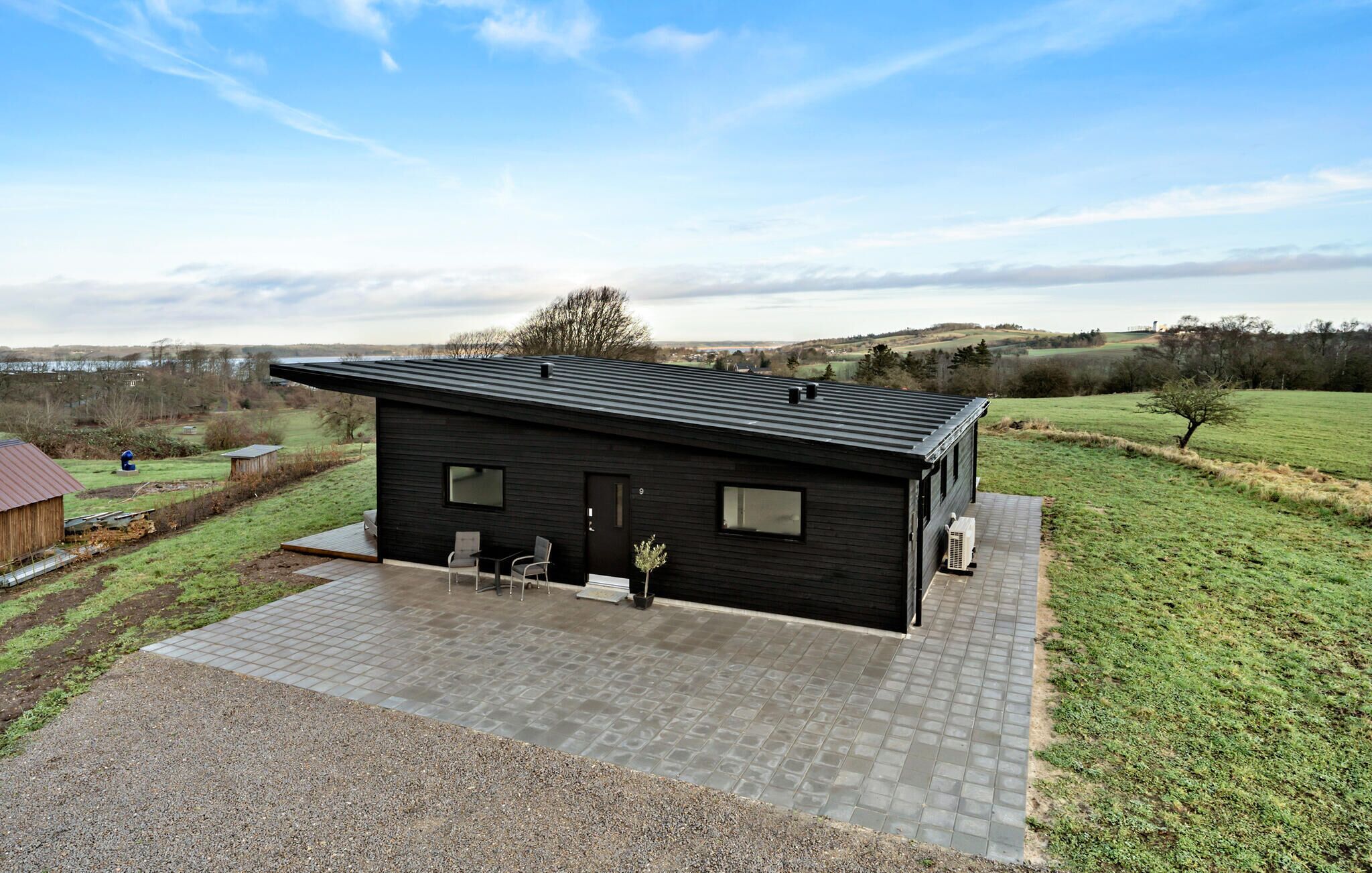 A modern black house with a flat roof, a paved driveway, and a small patio area with two chairs.