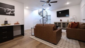 Ein modernes Wohnzimmer mit einem braunen Ledersofa, einem schwarzen Schreibtisch mit Schubladen, einer Wendeltreppe und einem flachbildfernseher an der Wand.